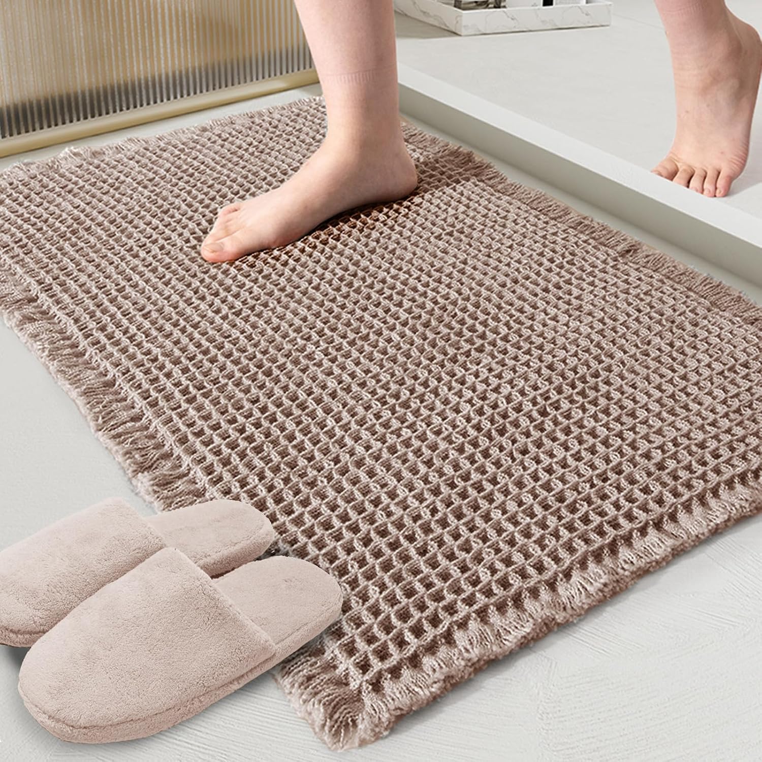 Tan waffle-texture bath mat with fringed edges on a bathroom floor next to slippers.