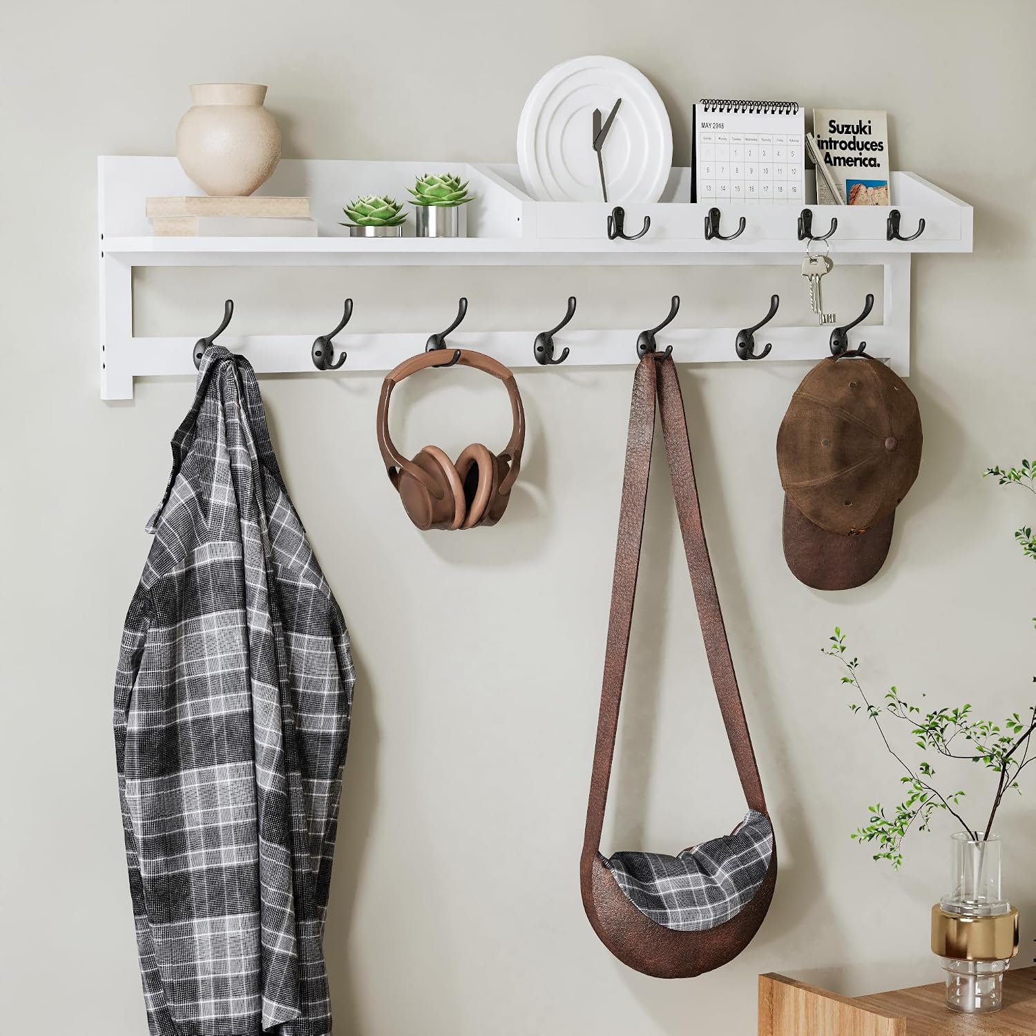 White wall mounted coat rack with a top shelf and rows of black dual hooks holding a jacket, headphones, a bag, and a cap.